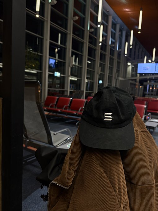 A black baseball cap with a white logo resting on top of a brown corduroy jacket, sitting on a seat in an empty airport terminal waiting area.