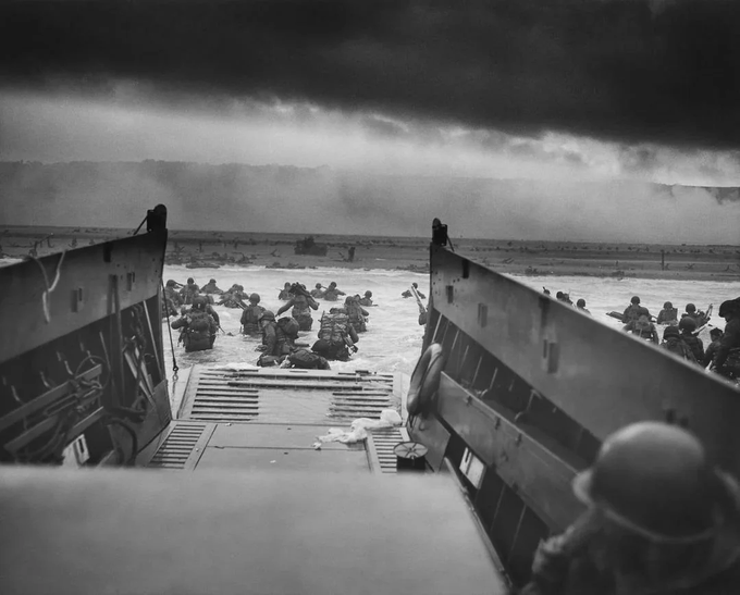 A historic black-and-white photograph from D-Day showing soldiers wading through water from a landing craft toward the beach at Normandy under a dramatic, cloudy sky.