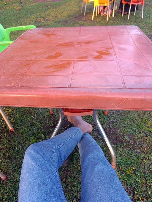 A first-person perspective photo looking down at a person's legs in blue jeans and bare feet extended under a reddish-brown plastic patio table on a grassy lawn.