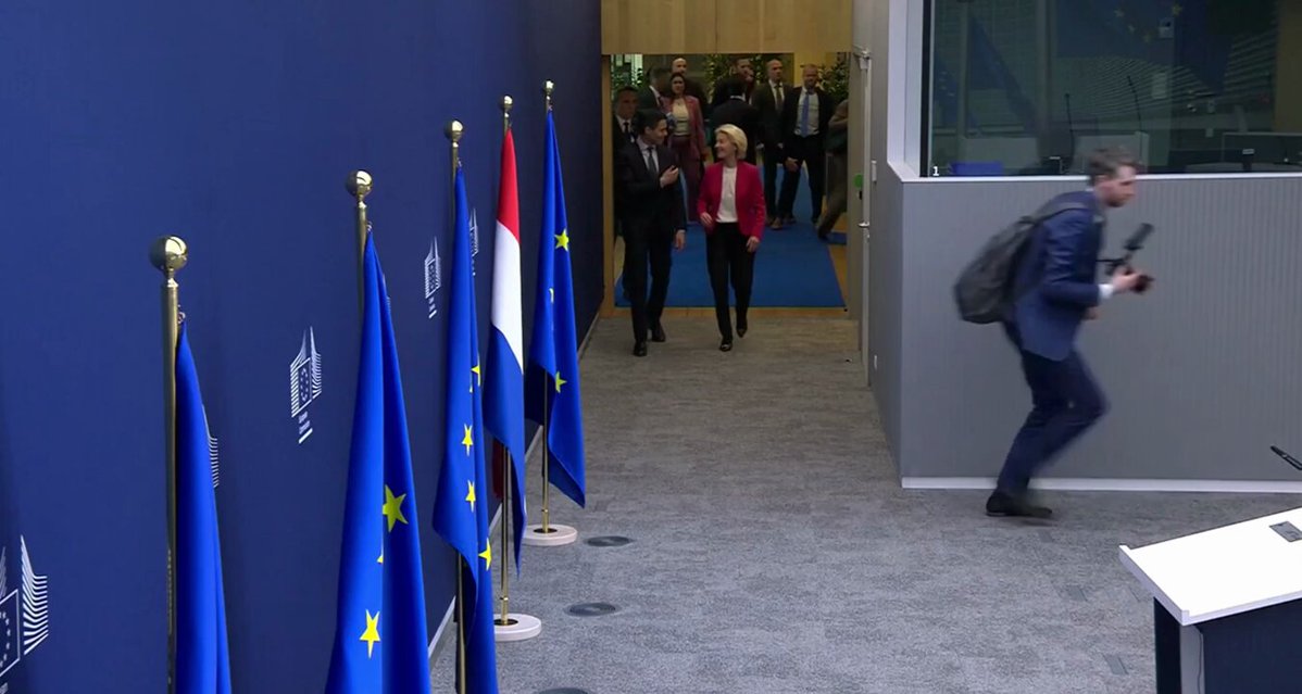 The image shows a group of people walking down an aisle with blue walls, surrounded by several flags including the European Union flag. One person appears to be running past them while another individual is carrying a backpack. The scene suggests that they are attending a meeting or event related to international affairs and finance.