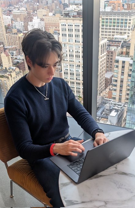 The image shows a young man sitting at a table using his laptop computer while wearing a blue sweater. The background features an urban cityscape with tall buildings, indicating that he might be working from home or in a co-working space located within the city. The caption of the image reads "At 19, I just left my dream job at @base A year ago, before joining, my life goal was to work at the frontier of crypto", suggesting that this young man is interested in and involved with cryptocurrency technology.