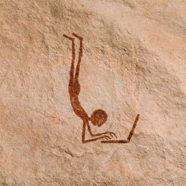 A photograph of an ancient rock painting (pictograph) showing a stick figure of a person in a handstand or diving pose, rendered in reddish-brown pigment on a textured stone surface.