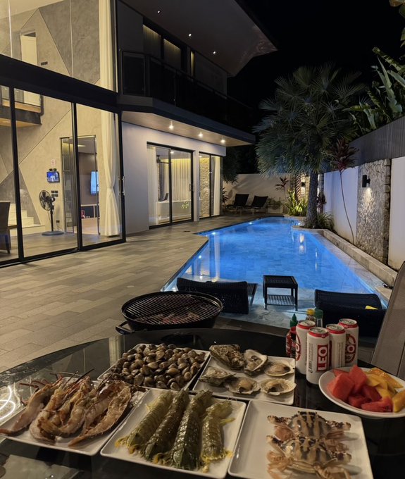 A nighttime view of a villa patio featuring a glowing swimming pool, a grill, Leo beer, and plates of seafood including shrimp, fish, oysters, and crab ready for a meal.