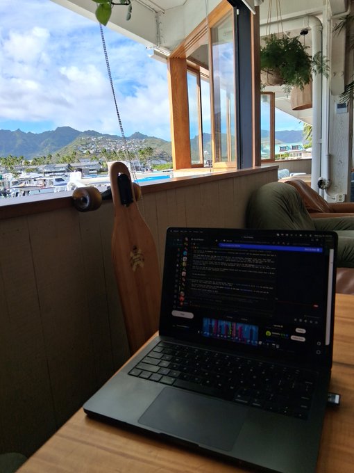 A laptop sits open on a wooden table by a window, displaying a chat application, with a scenic view of mountains, harbor, and boats visible outside, along with a wooden longboard skateboard leaning against the wall.