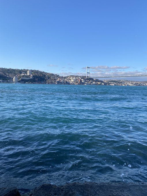 A scenic view of the Bosphorus strait in Istanbul, showing blue water with a suspension bridge and hilly shoreline under a clear sky.