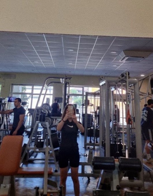 A person taking a mirror selfie with their phone in a gym, surrounded by weight machines and other people working out in the background.