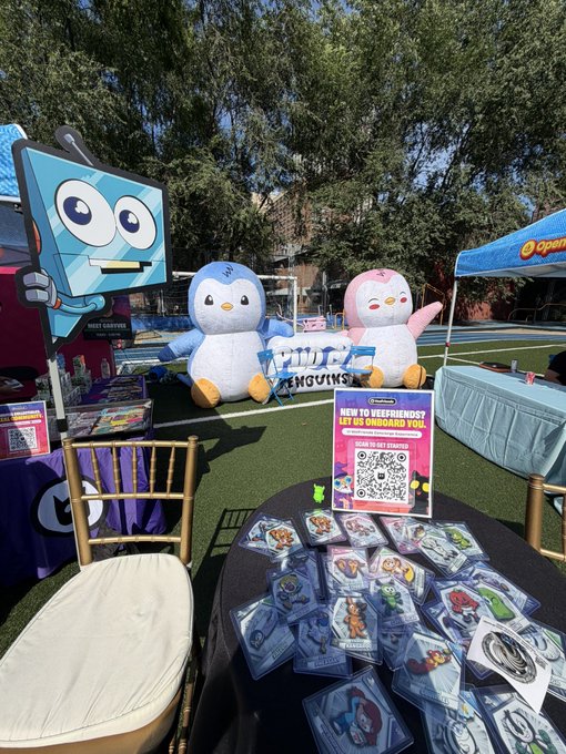 An outdoor booth displaying merchandise for 'Keeblings' alongside a large blue robot character, pink and blue penguin mascots, and a sign encouraging attendees to 'SWEEP TO SEE CREATIVES' via a QR code.