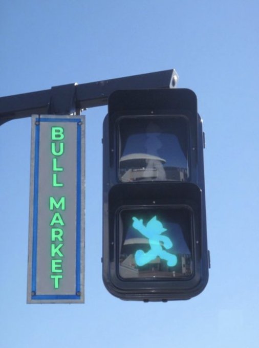 A traffic light showing a green pedestrian crossing symbol of a walking figure, mounted next to a vertical illuminated 'BULL MARKET' street sign against a blue sky.