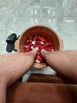 A first-person view shows someone sitting with their feet in a wooden tub filled with water and red rose petals, with a black slide sandal visible on the tiled floor nearby.