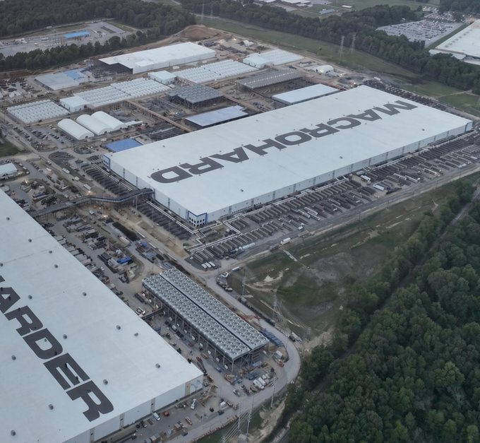 Aerial view of a large industrial facility with "MACROHARD" painted on the roof of the main building, surrounded by parking lots and forested areas.