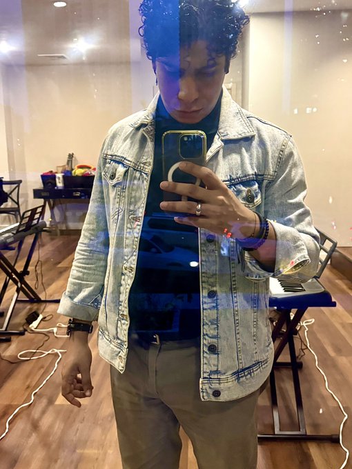 A mirror selfie of a young man with curly hair wearing a light denim jacket and dark shirt, holding a phone and making a peace sign in what appears to be a music studio with a keyboard.