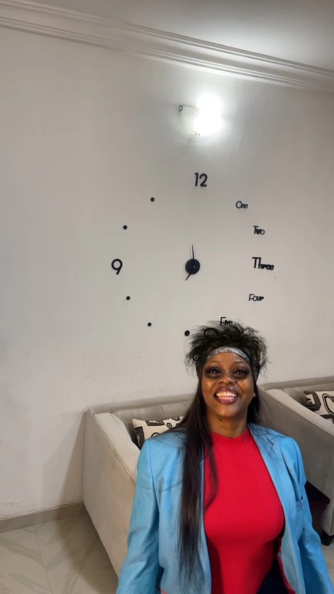 A smiling woman in a red top and blue blazer poses indoors beneath a large wall clock with abstract number placements.