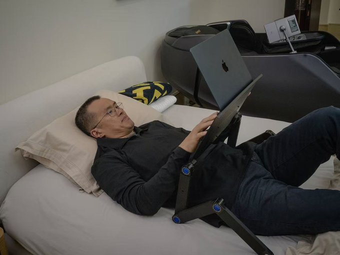 A man wearing glasses reclines on a light-colored couch, using a laptop placed on an adjustable lap stand.