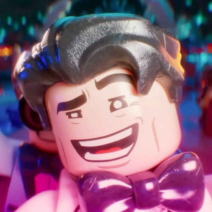 A close-up of a Lego figure depicting a smiling man with dark hair wearing a purple bow tie, set against a colorful blurred background.