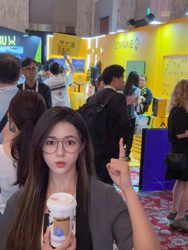 A young woman with glasses holds a coffee cup and points upward in a crowded, brightly lit indoor venue, with 'K11' visible on a sign in the background.