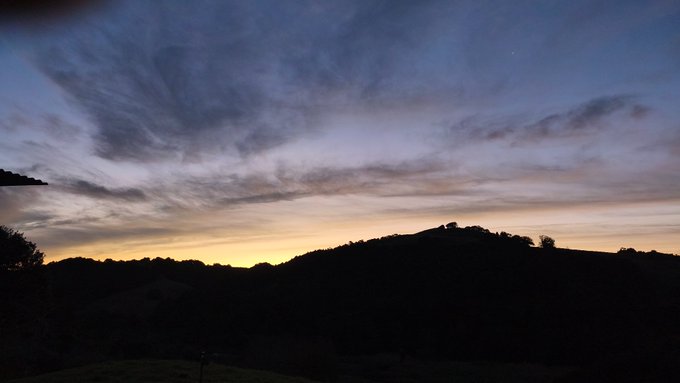 A dusk landscape photo showing a silhouetted hillside beneath a vast sky with wispy blue clouds and a warm orange glow along the horizon.
