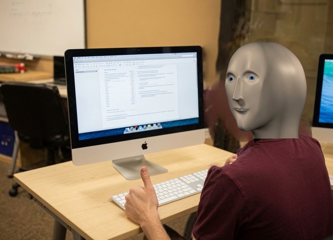 A person wearing a gray robot mask sits at a desk working on an Apple iMac displaying a spreadsheet, in what appears to be an office setting.