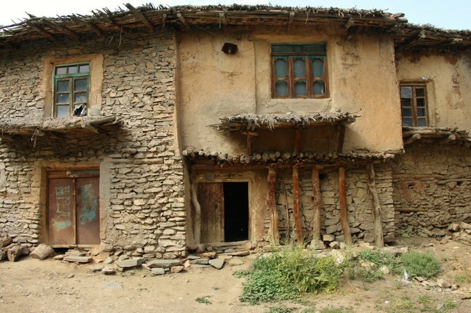 A rustic two-story mud and stone house features weathered earthen walls, wooden-framed windows, a thatched roof, and a propped-up wooden support beam, exemplifying traditional rural architecture.

This image directly illustrates the "Rain and Mud Houses" cultural photography piece, showcasing traditional earthen construction techniques that reflect local building heritage and the textures of vernacular architecture.