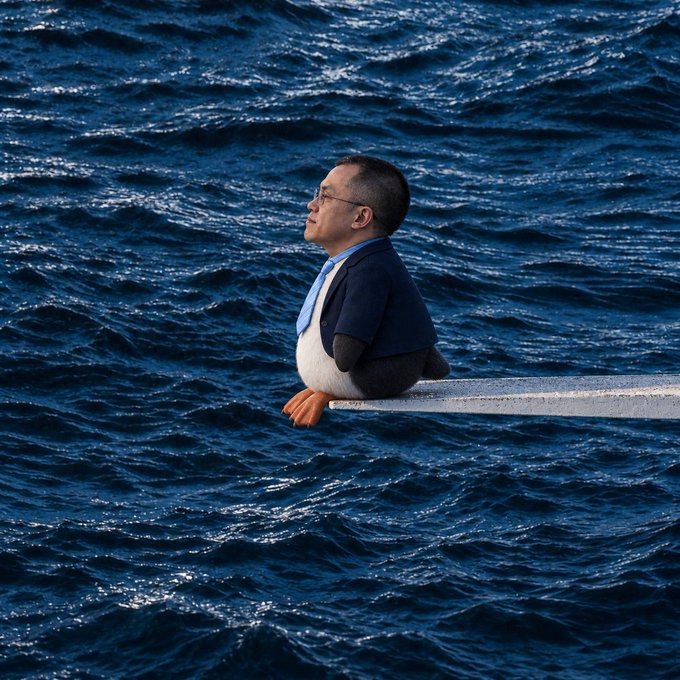 An AI-generated image shows a penguin with a man's head (wearing glasses and a suit jacket) sitting on a railing overlooking a vast blue ocean.