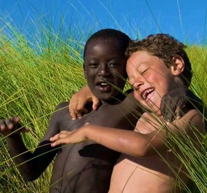 Two young boys laughing and hugging while partially hidden in tall green grass against a clear blue sky.