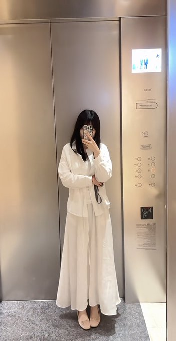 A woman in an elegant white blouse and flowing white skirt takes a mirror selfie inside an elevator, holding up her phone to capture the reflection. The accompanying post suggests a "good morning" greeting tied to the market opening, though no financial text or numbers are visible in the image itself—only the elevator's modern metallic interior and button panel.