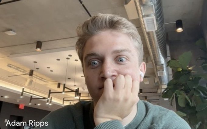 A young man with blonde hair and wide eyes covers his mouth in surprise in what appears to be a cafe or office setting, with text identifying him as 'Adam Ripps' in the corner.