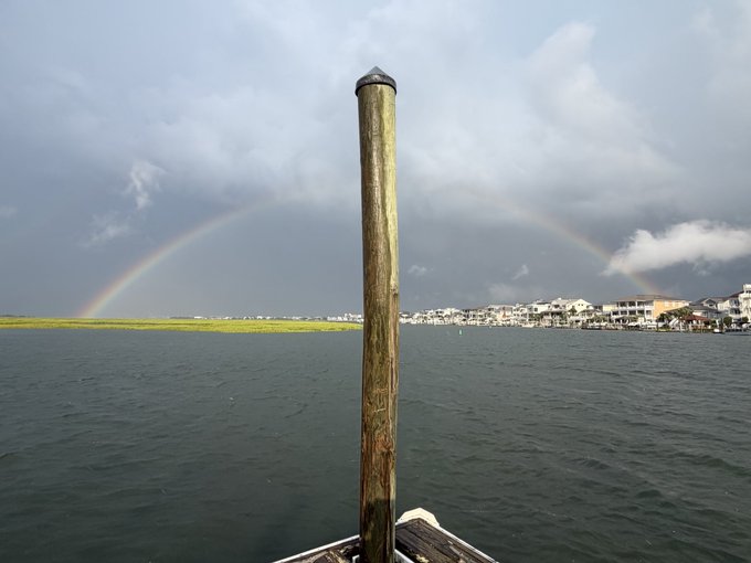 A photo taken from a boat showing a wooden piling in the foreground with a rainbow arching across the cloudy sky over a body of water and distant shoreline houses.