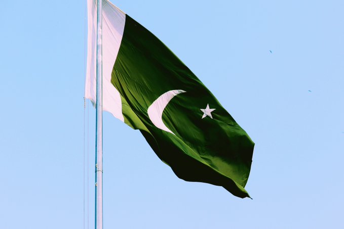 The national flag of Pakistan flies against a clear blue sky, featuring its distinctive dark green field with a white crescent and star, alongside a white vertical stripe at the hoist. The image accompanies news of Pakistan's crypto regulator pursuing technical and Shariah compliance reviews of digital assets, following an Islamic scholar's ruling against crypto payments.