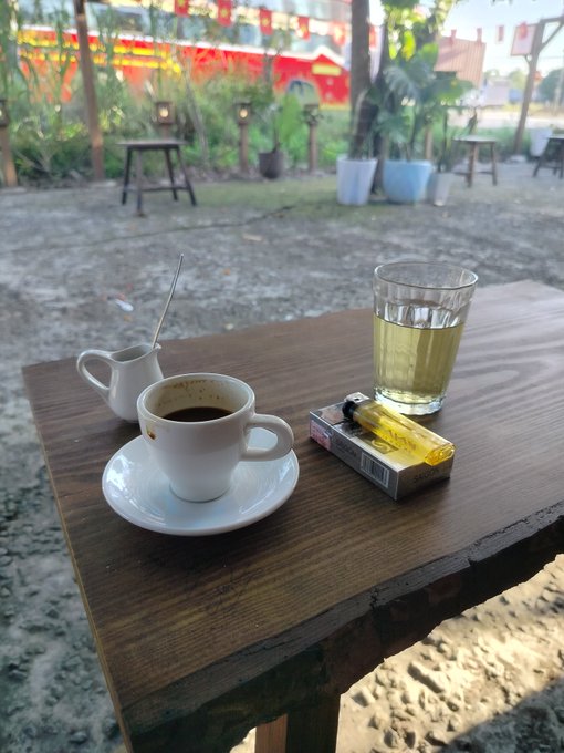 A small cup of black coffee and a glass of tea sit on a wooden table at what appears to be a beachside cafe, accompanied by a pack of cigarettes. The relaxed scene contrasts with the post's somber tone about portfolio losses during the Robinhood Summit, evoking a sense of someone seeking solace in simple comforts amid financial disappointment.