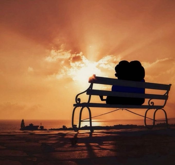 A silhouette of a couple sitting together on a park bench at sunset, watching the sun set over the ocean with golden light rays streaming through clouds.