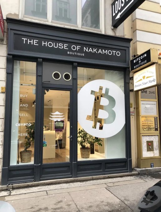 A storefront called 'THE HOUSE OF NAKAMOTO BOUTIQUE' featuring a large Bitcoin 'B' logo in the window and signs reading 'BUY AND SELL CRYPTO HERE' and 'BITCOIN BOUTIQUE'.