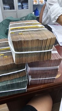 Multiple thick stacks of banknotes bundled together sit on a wooden table, with a person in a white shirt partially visible in the background.