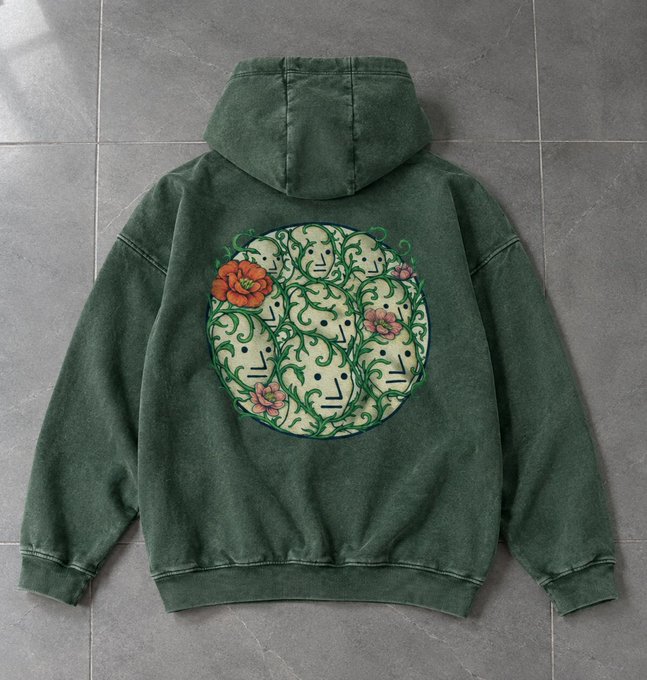 A faded green hoodie lies on a tiled floor, featuring a large circular back graphic with intertwined faces, vines, and red and pink flowers.