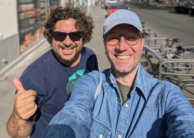Two smiling men taking a selfie outdoors on a sunny city sidewalk; the man on the left wears a blue t-shirt and sunglasses, while the man on the right wears a denim shirt, glasses, and a light blue cap.