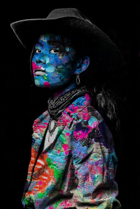 A portrait of a child in a black cowboy hat and bandana, with face and clothing covered in vibrant neon paint splatters against a dark background.