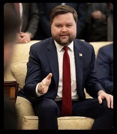 A formal photograph of a bearded man in a dark blue suit with a red tie, seated on a beige chair and gesturing with his hand, with a small flag pin on his lapel.