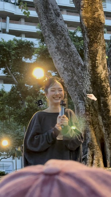 A young woman in a dark sweater smiles while holding a microphone outdoors at dusk, standing near a large tree with a building visible in the background.