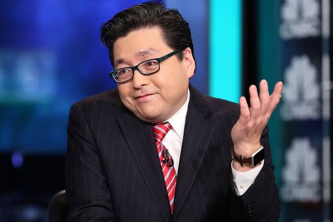 A man wearing glasses, a dark pinstripe suit, and a red striped tie gestures with his hand while appearing on a television set with blue lighting.