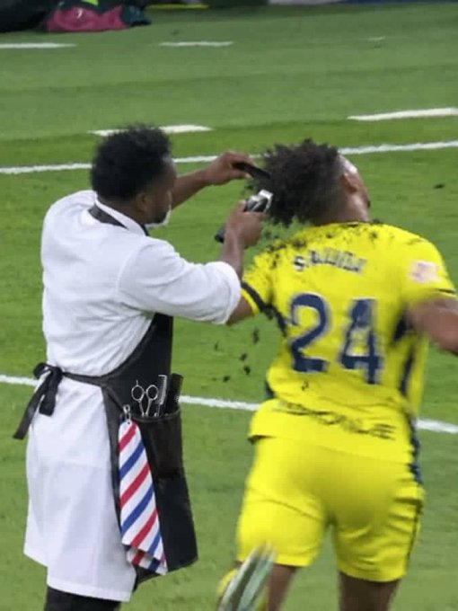 A player wearing a yellow jersey with "S. AKIMOV" and the number "24" gets a haircut from a barber on the field, supporting the claim that "Vini Jr. did nothing wrong" by highlighting a humorous moment of unprofessional behavior during a match.
