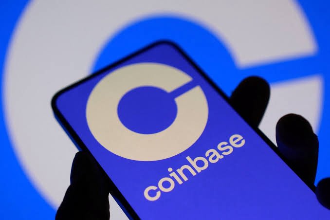 A hand holds a smartphone displaying the Coinbase logo, with the company's name visible beneath it against a blurred blue background. The image accompanies news of Coinbase's disappointing Q2 results, highlighting the company's financial struggles amid weaker crypto prices and reduced trading activity.