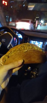 A hand holding a piece of bread or pastry inside a car at night, with the dashboard and street lights visible in the background.