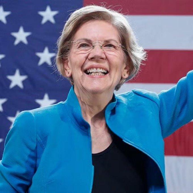 Senator Elizabeth Warren smiles and waves, wearing a bright blue jacket with an American flag in the background.