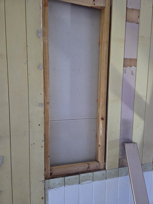 A construction site shows a rough wooden-framed window opening in a wall with yellow paneling, revealing gray sheathing behind it, with a loose board leaning against the siding below.