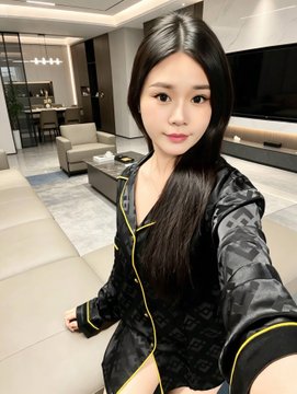 The image features a young woman with long hair wearing a black shirt, sitting on a couch in a living room setting. She appears to be posing for a photo or taking part in an event related to the post she has just posted. The background of the image shows a dining table and chairs, suggesting that this gathering might be taking place at someone's home.