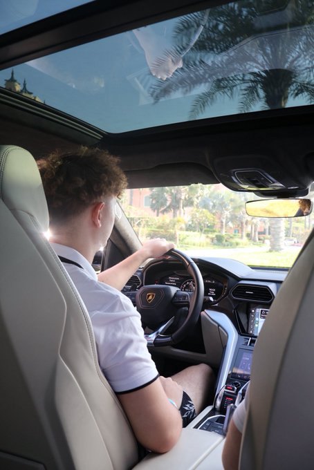The image shows a young man with curly hair driving a Lamborghini, gripping the steering wheel which displays the brand's iconic bull logo, while palm trees and a sunny landscape roll by through the windshield and panoramic sunroof. The "GM GM GM" caption likely expresses envy or excitement, contrasting the driver's apparent luxury lifestyle with the poster's own situation.