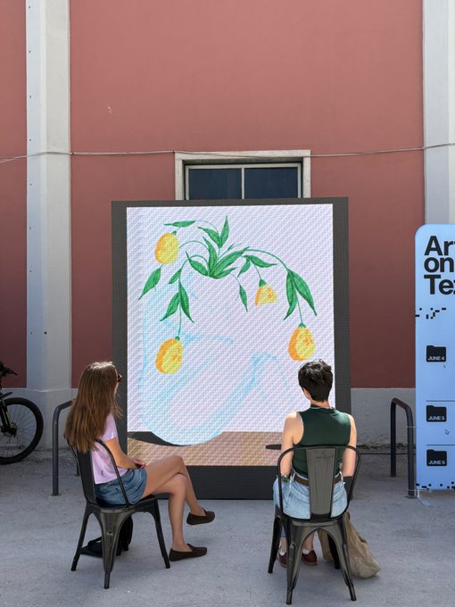 The image shows two people sitting at a table, looking at a large painting of lemons hanging on a wall behind them. They are positioned near a window that overlooks a building with pink walls. The scene appears to be set up for an art event or exhibition, as there is also a sign in the background that reads "Ar on te".