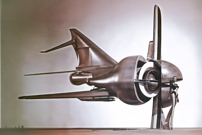 A sleek, metallic futuristic aircraft or vehicle sculpture with sharp, angular wings and a rounded central body, displayed on a wooden surface.