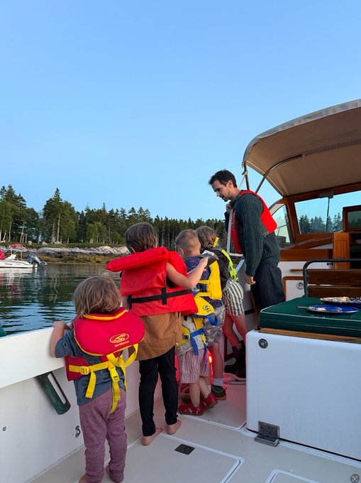 A family of four children in life jackets stand along the railing of a boat in a Maine harbor, accompanied by an adult man, looking out at the water and tree-lined shore.