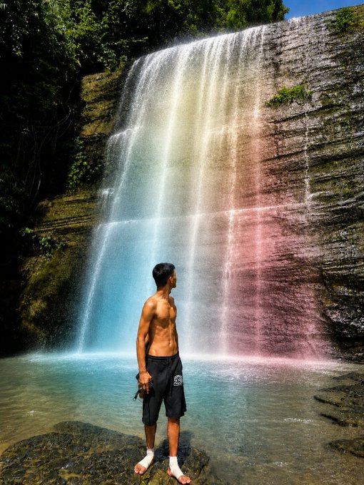 A shirtless man stands on rocks in front of a waterfall that is illuminated with rainbow colors.