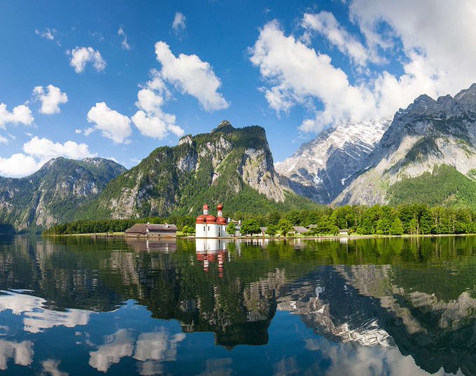 A scenic landscape featuring a small church with twin red-domed towers on the shore of a calm lake, surrounded by lush green mountains under a blue sky with fluffy white clouds.